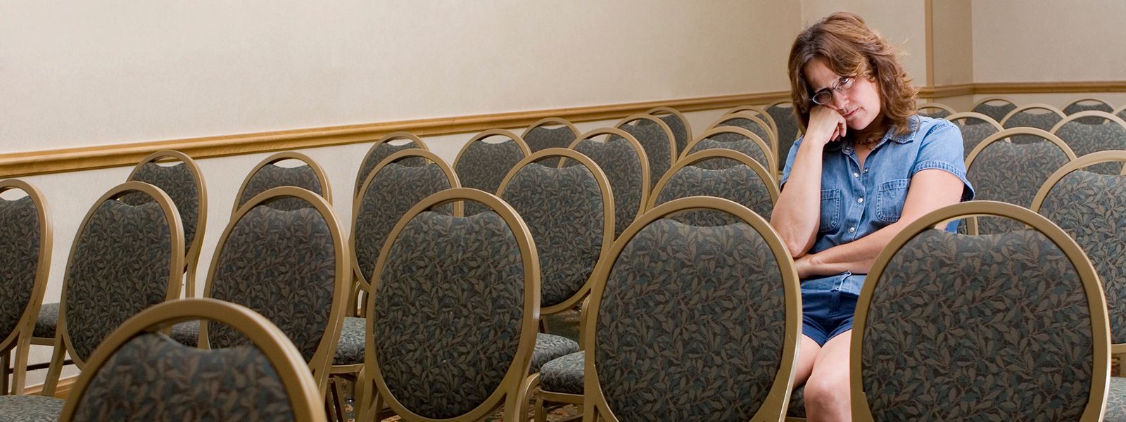 A woman sits alone among rows of empty chairs in a quiet conference or meeting room, resting her head on her hand and appearing thoughtful or concerned.