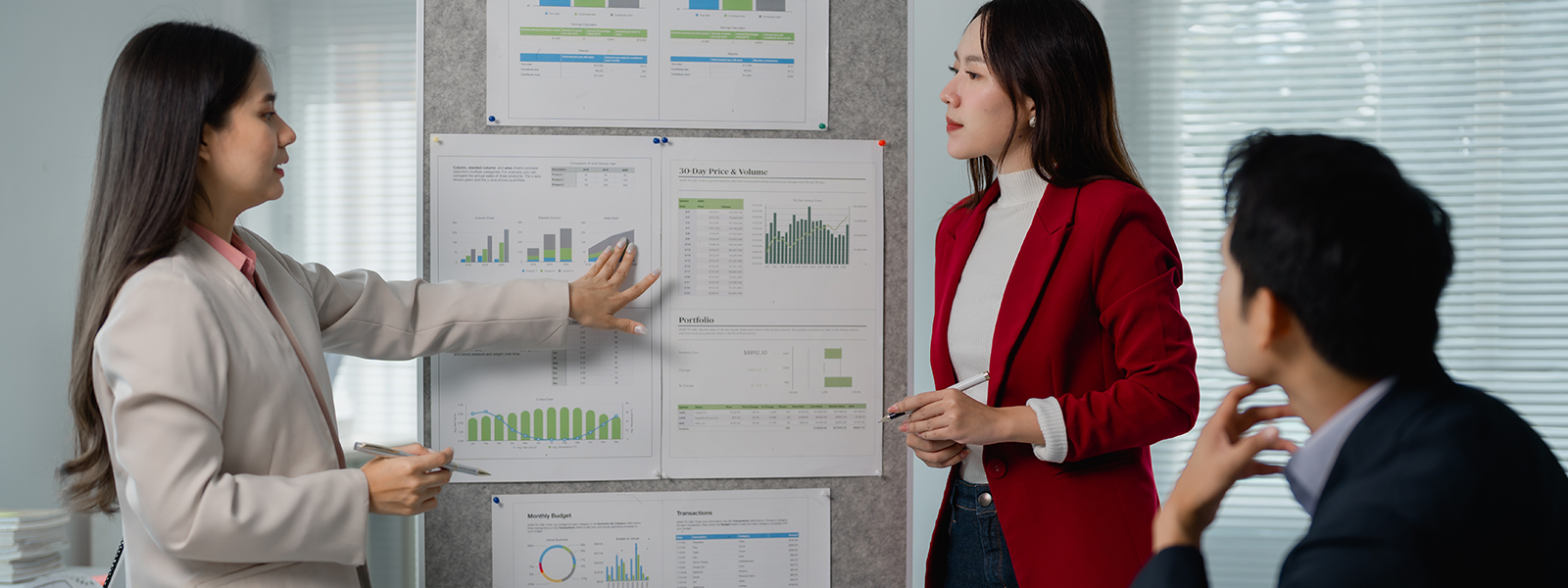 Three colleagues in a modern office review printed charts on a bulletin board, with one person pointing to bar and line graphs while others listen and discuss performance data.