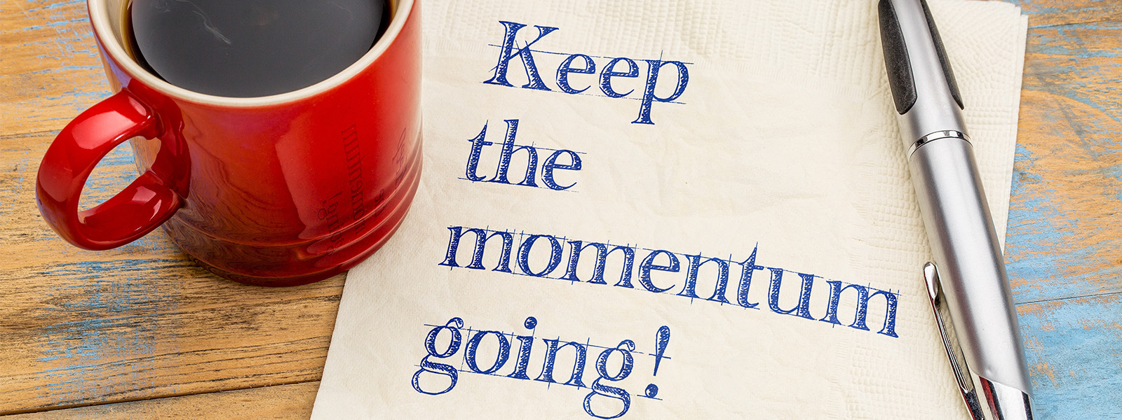 Text on piece of paper saying KEEP THE MOMENTUM GOING, with a coffee mug and pen on side