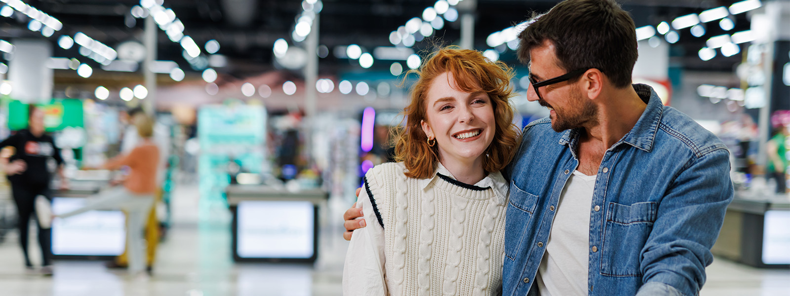 A happy couple walks through a brightly lit shopping mall. The woman, wearing a white sweater, smiles while looking forward, while the man, wearing a denim jacket and glasses, looks at her with a smile.
