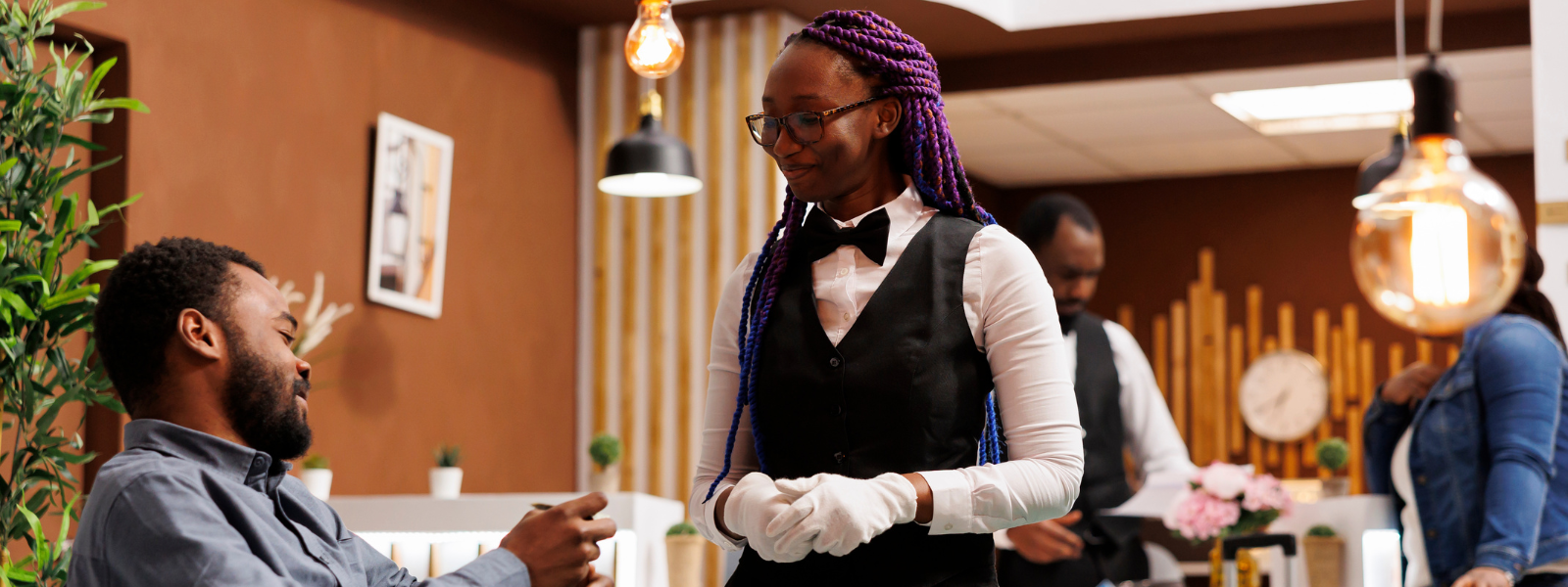 Smiling waiter in a vest and bow tie interacting warmly with a seated guest in a cozy, modern restaurant setting.